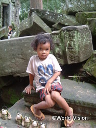 Young child sits on mossy, ancient stone blocks in a ruin, with small penguin figurines lined up nearby (World Vision watermark).