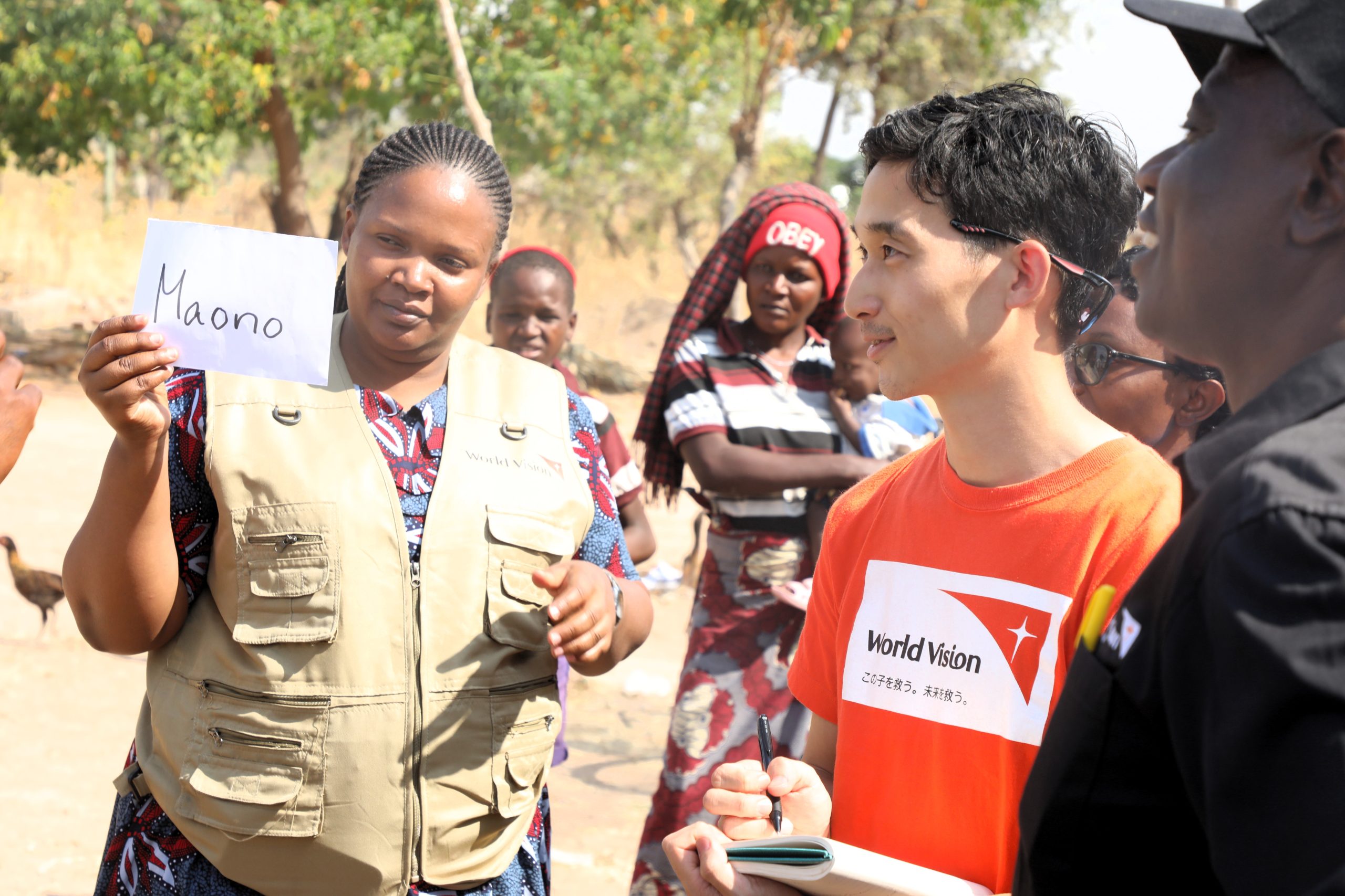Woman in a tan utility vest holds up a white card with the word 'Maono' while a man in an orange World Vision shirt watches, in an outdoor gathering with onlookers and trees in the background.