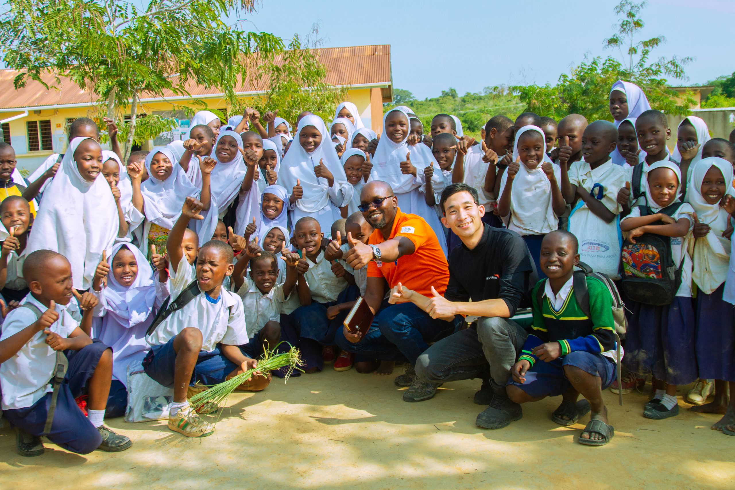 Large group of smiling students in uniforms posing outdoors at a school, with two men in front giving thumbs up.