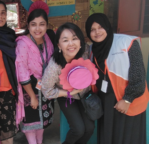 Four women posing together at a community event; the front woman holds a pink hat with ribbons while smiling at the camera.