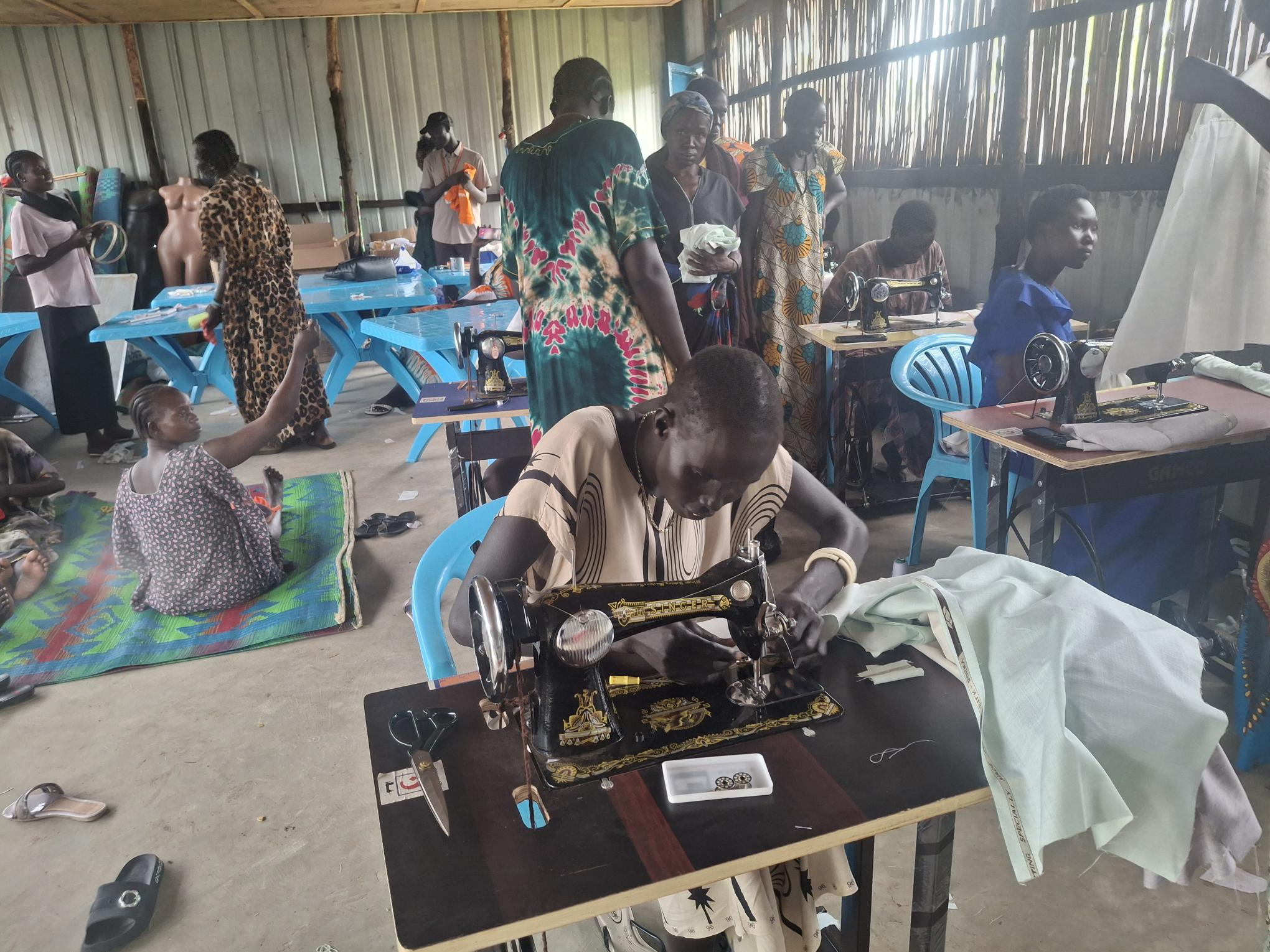 Group of people in a workshop with sewing machines and fabric on blue tables, inside a shed-like space.