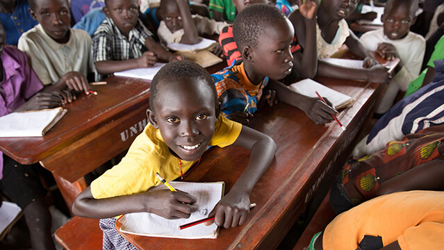 木製の机で学ぶ男子児童が笑顔を向ける。教育支援により地域の子どもたちが未来への力を育む様子。