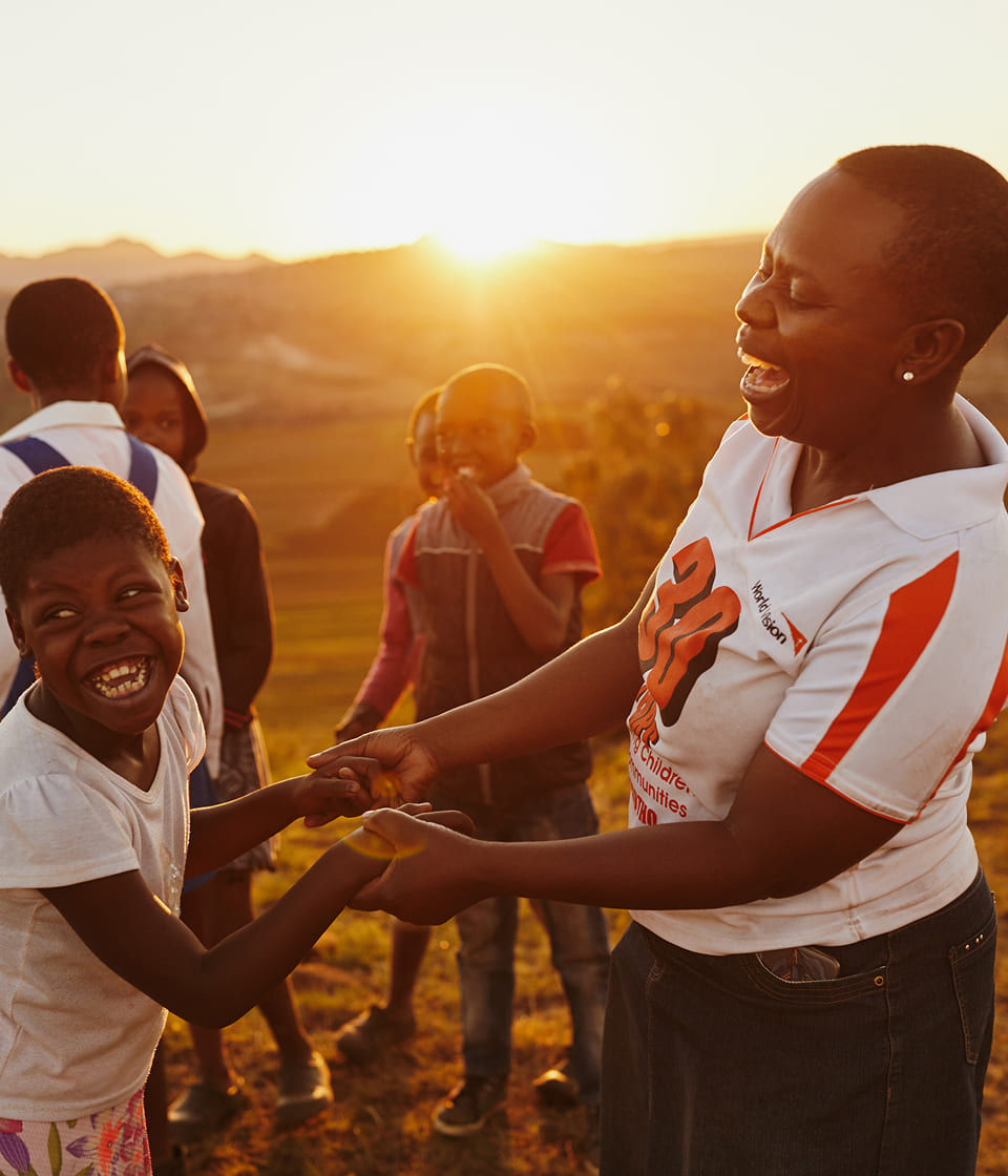 夕日を背景に、アフリカの村で子どもと大人が笑顔で手を取り合い、地域支援の喜びを分かち合う様子。