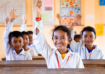 明るい教室で学ぶ子どもたちが手を挙げる姿。企業による教育支援が未来を照らす。