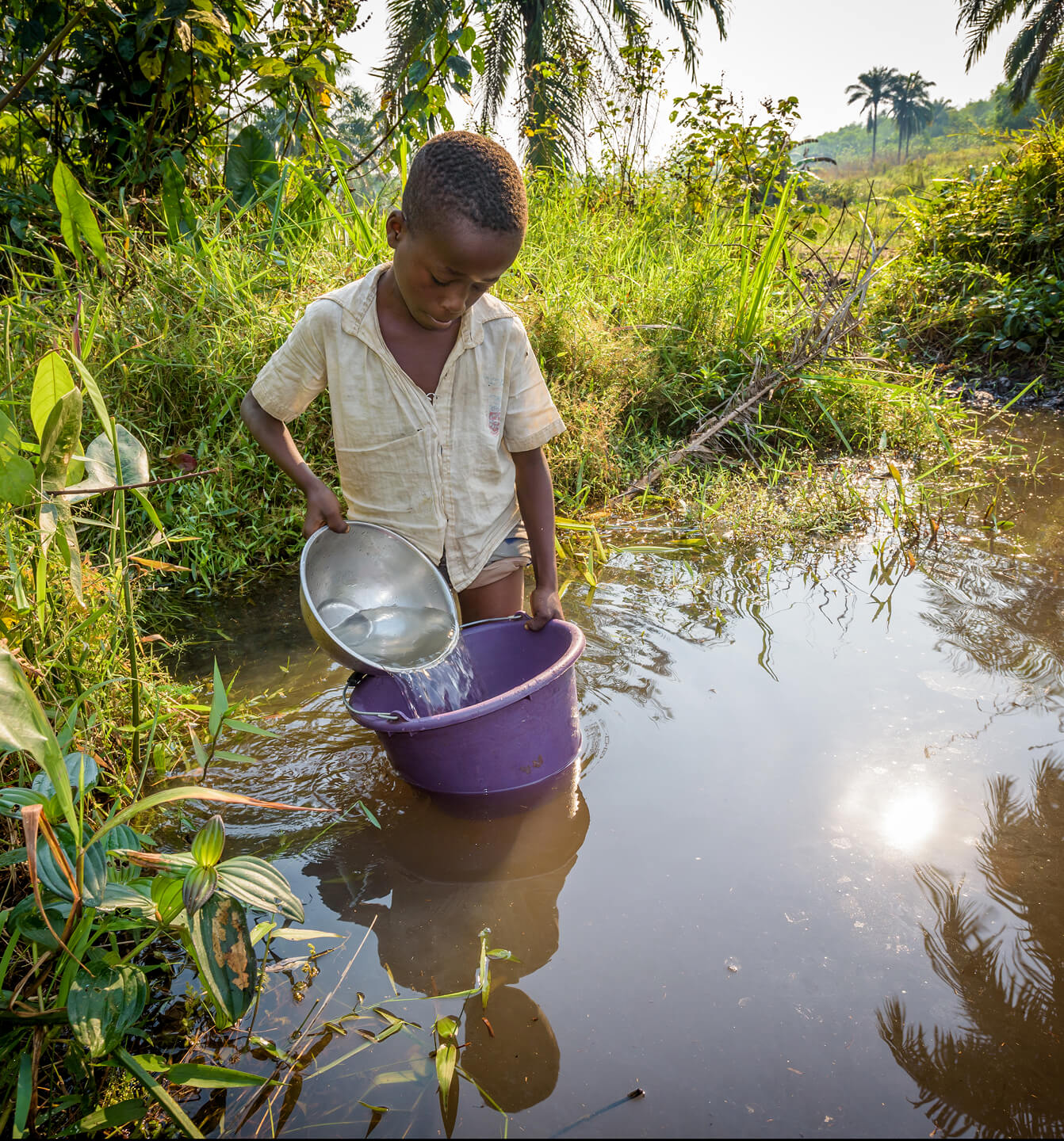 水辺で生活用水を集める少年。安全な水を得るための支援が、子どもたちの健康と未来を守る重要な活動であることを伝える。
