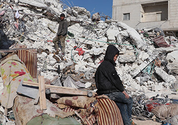 瓦礫に囲まれた被災地で途方に暮れる子どもと住民。地震被害からの復旧と人道支援の重要性を伝える場面。