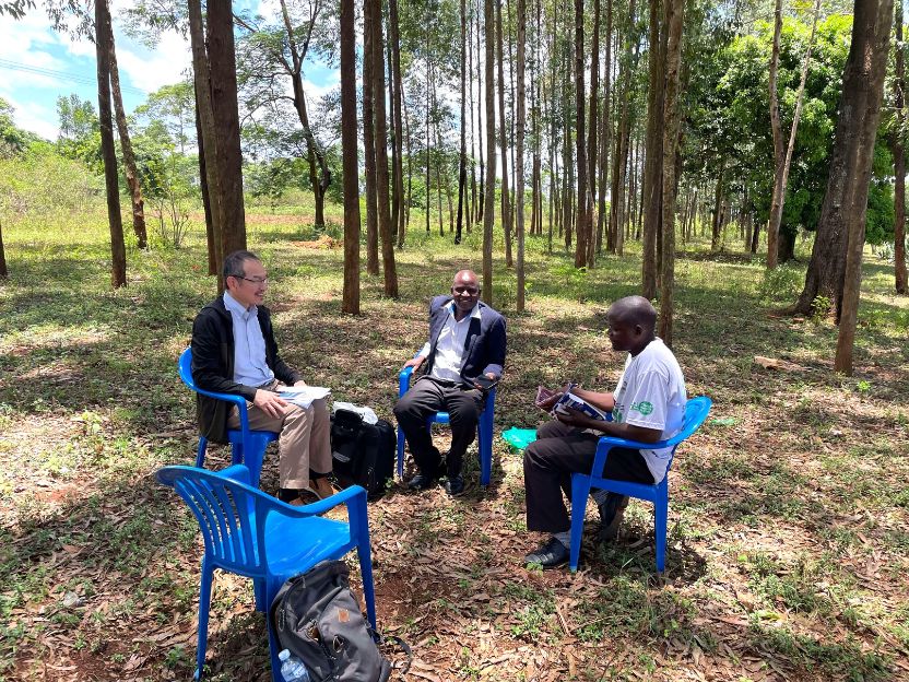 Three men sit on blue plastic chairs in a forest, having a discussion outdoors. One holds papers, another smiles, the third reads a book.