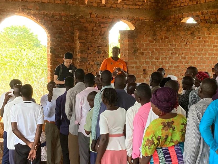 Group of people lined up indoors in a brick building, two men at the front supervising a distribution or registration event.