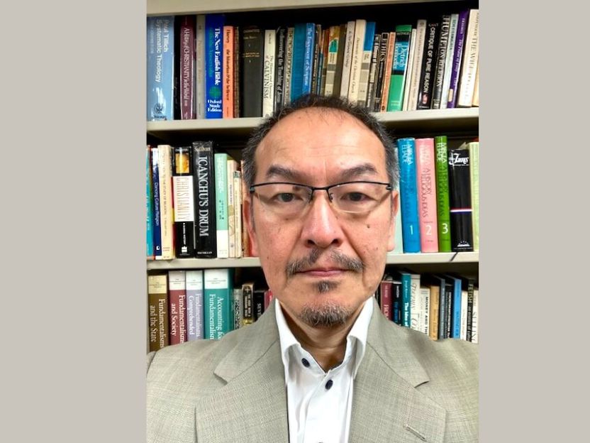 Portrait of a middle-aged man with glasses in a light suit, standing in front of a bookshelf filled with colorful books.