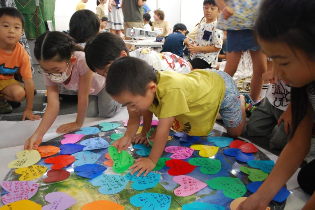 Children gather around a large circular map, placing colorful heart-shaped notes on its surface in a classroom activity.