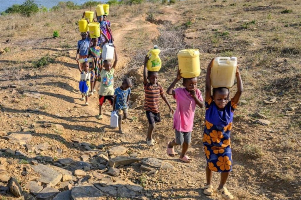 Group of children walking in a dry, rocky landscape carrying yellow water jugs on their heads in a line.