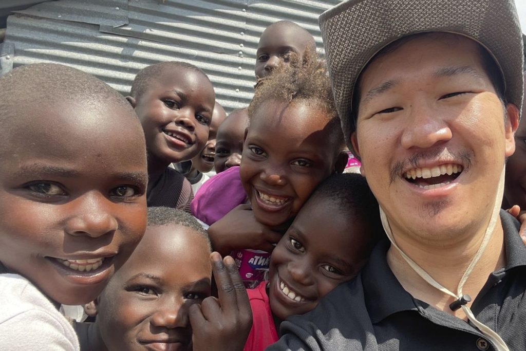 Smiling Asian man wearing a hat takes a selfie with a joyful group of African children close around him.
