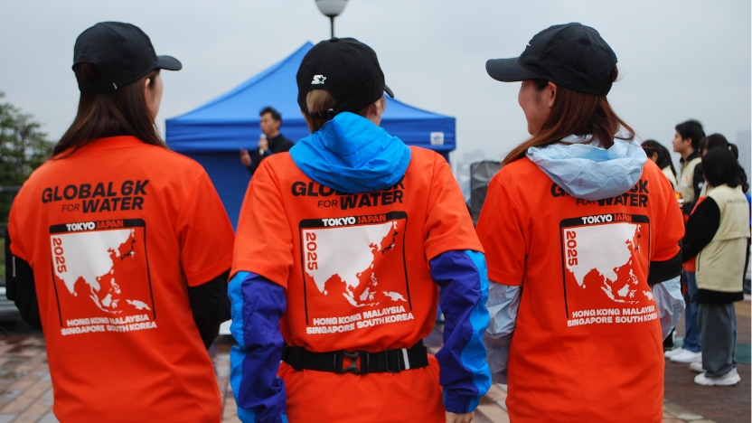Three volunteers wearing orange 'Global 6K for Water' shirts stand with their backs to the camera at an outdoor event, blue tents visible in the background.