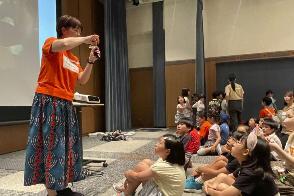 Presenter in an orange shirt and patterned skirt speaks into a microphone to a group of children seated on the floor in an auditorium.