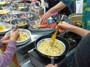 People cooking at a station: hands mixing chopped vegetables in aluminum bowls with bags of snack mix nearby on a metal countertop.