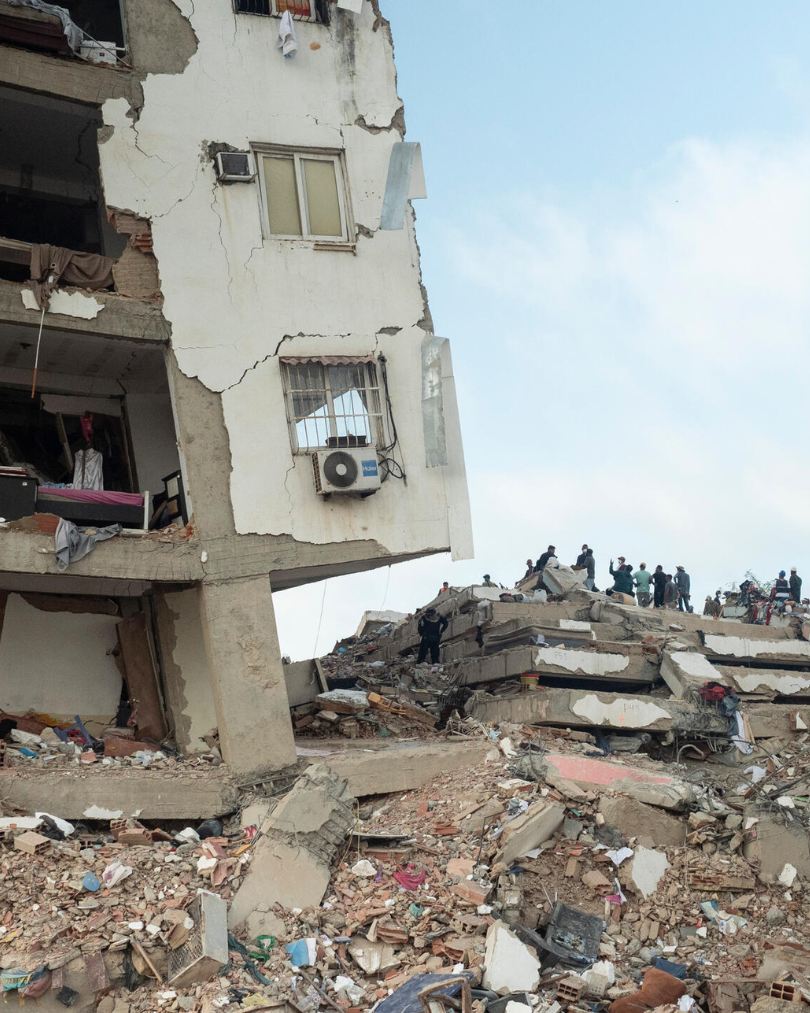 Collapsed multi-story building with rubble and debris; rescue workers and onlookers on a ruined terrace, disaster zone scene