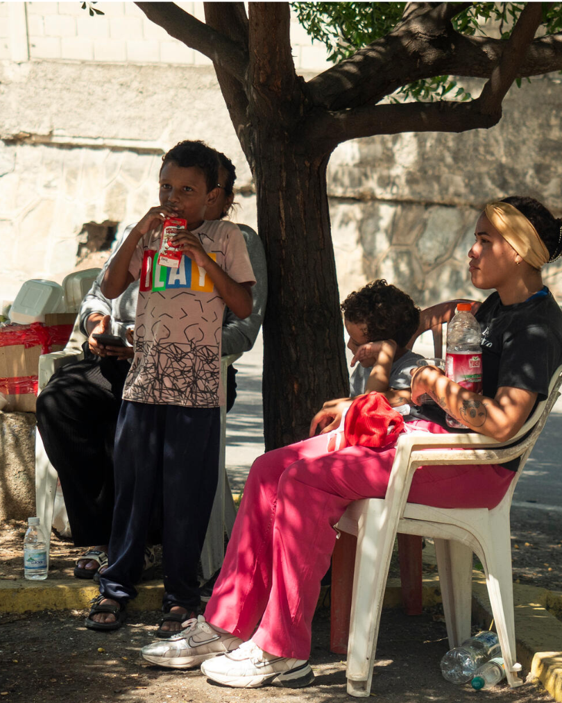 A child standing and drinking from a juice box while others rest on plastic chairs under a tree in a shaded outdoor area.