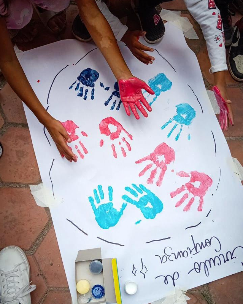 Children's hands covered in red, pink, blue paint pressed onto a large white poster, forming colorful handprints inside a circle drawn on the ground, art supplies nearby.