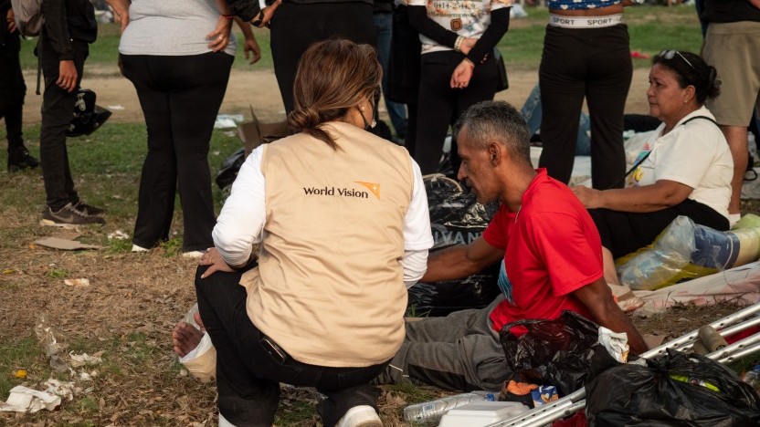 Volunteer from World Vision kneels beside a man in a red shirt on a grassy area, offering assistance among bags and debris at a relief scene.