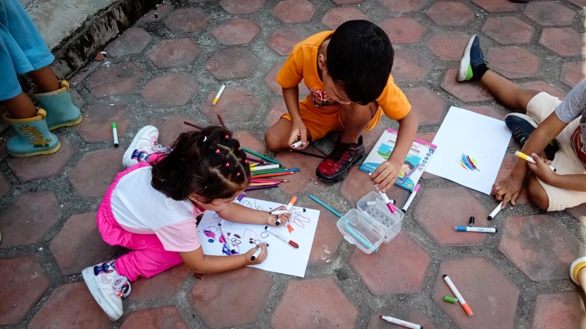 Three children sit on a tiled outdoor surface, drawing on papers with colored markers and pencils, surrounded by art supplies.