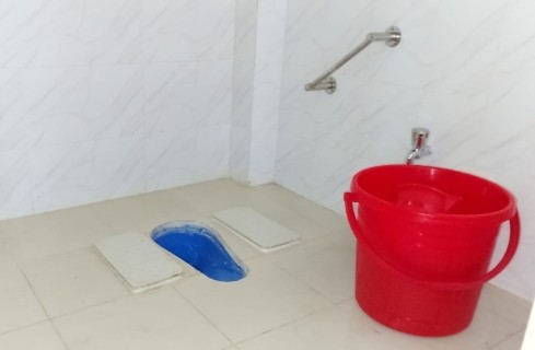 Public bathroom featuring a blue squat toilet with two white platform covers pulled aside, a red bucket of water nearby, and tiled walls and floor.