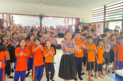 Group of schoolchildren in orange vests standing together and making hand gestures in a classroom hall.