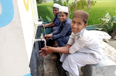 Four boys in traditional clothing at a wall-mounted water tap, washing hands outdoors near green fields.