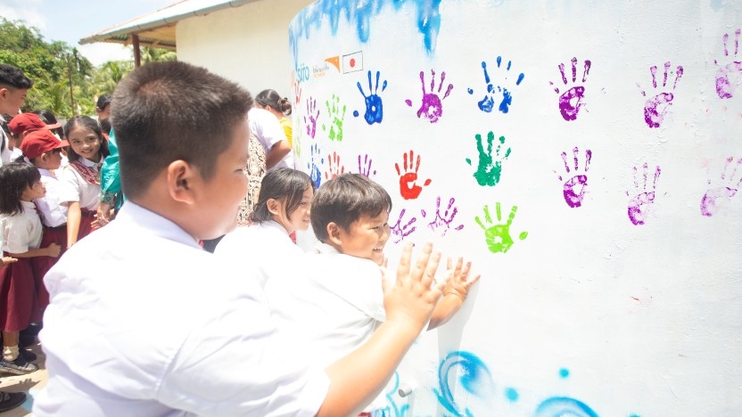 Children in white shirts press colorful handprints onto a white wall mural during a school activity.