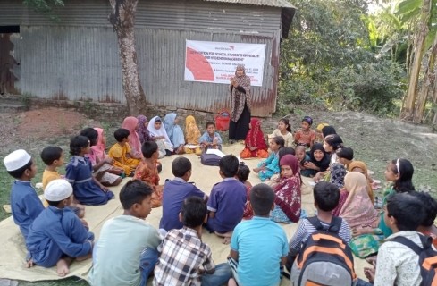 Woman standing and addressing a circle of children seated on mats outdoors, with a banner in the background.