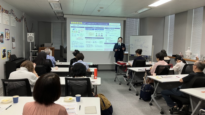Lecturer in a suit presenting to adults in a conference room, with a slide projected on a screen and a whiteboard nearby.