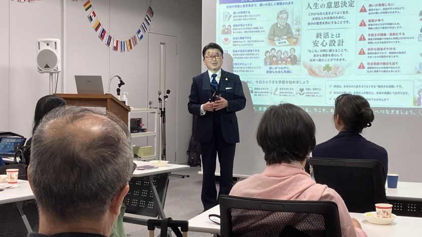 Man in a dark suit giving a talk to an audience in a conference room, with a projection screen showing slides in Japanese behind him.