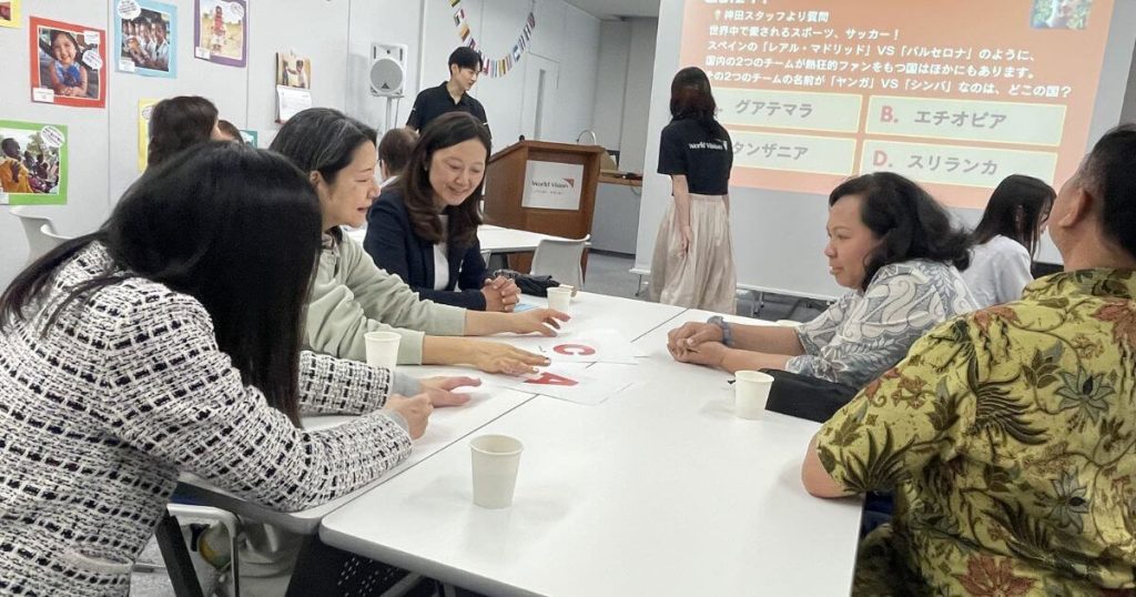 A group of adults sits around a white table in a classroom, collaborating on papers with red letters during a workshop, cups on the table with a projector screen in the background.