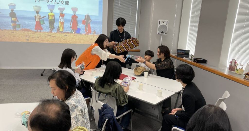 Group of adults and children seated around a long table in a meeting room, exchanging gifts during a community workshop.