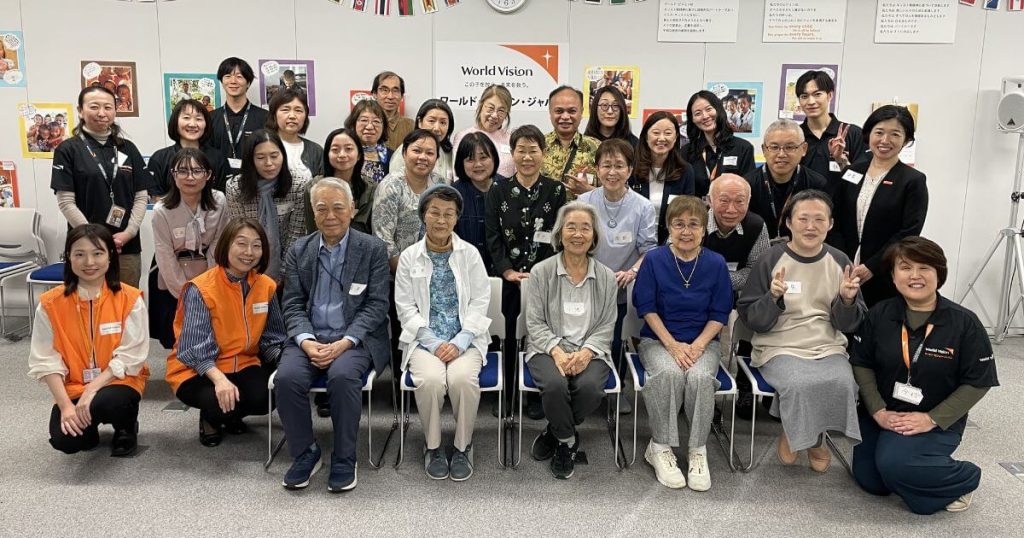 Group photo of about 30 volunteers and staff posing in a World Vision event room with a banner in the background.