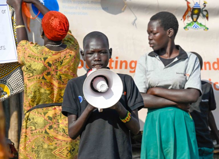 Boy with a megaphone addressing a crowd at an outdoor event, with a woman in colorful dress and a man in a grey shirt nearby.