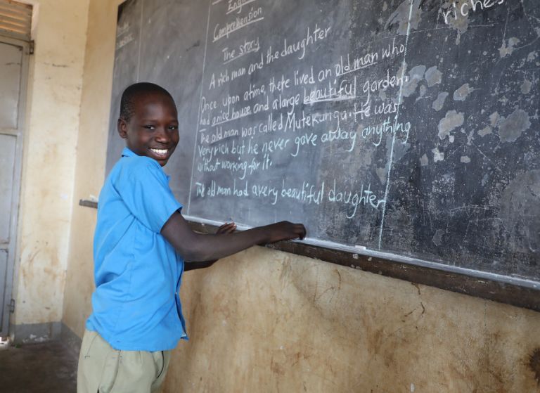 Young boy in a blue shirt smiles at the camera while writing on a chalkboard in a classroom.