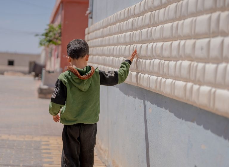 Young boy in a green hoodie touches a light-colored textured wall while standing on a sunny sidewalk.