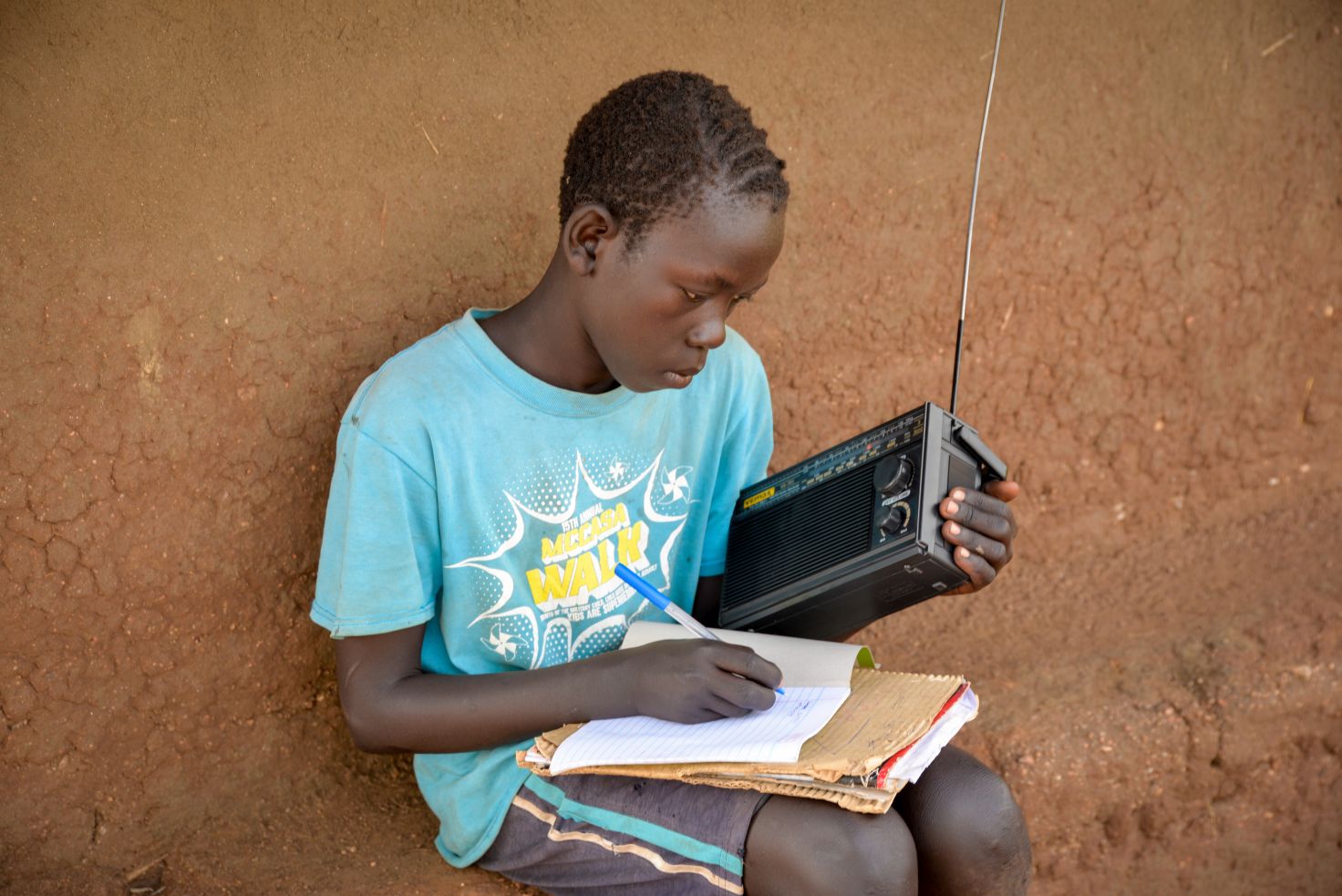 Boy sitting against a dirt wall, writing in a notebook while holding a portable radio.