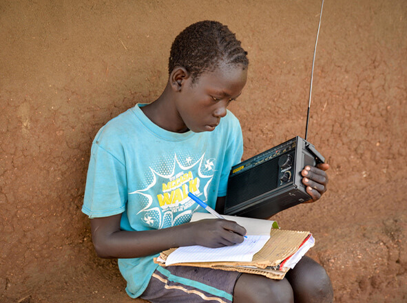 Boy sitting against a brown clay wall, writing in a notebook with a pen while holding a portable radio beside him on his lap