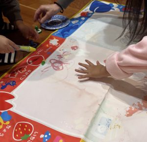 Children's hands drawing with markers on a colorful activity mat, with a blue plate nearby and paint marks around.