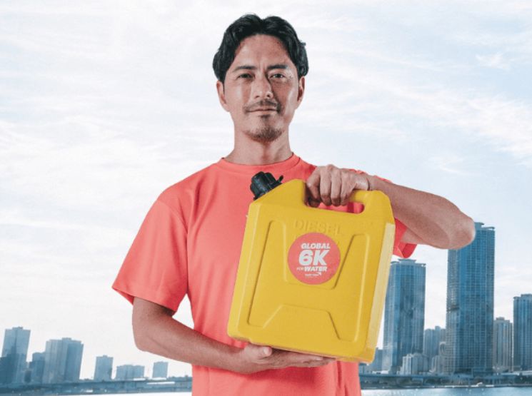 Man in a red shirt holding a yellow water container labeled 'Global 6K Water' with a city skyline in the background, outdoors.
