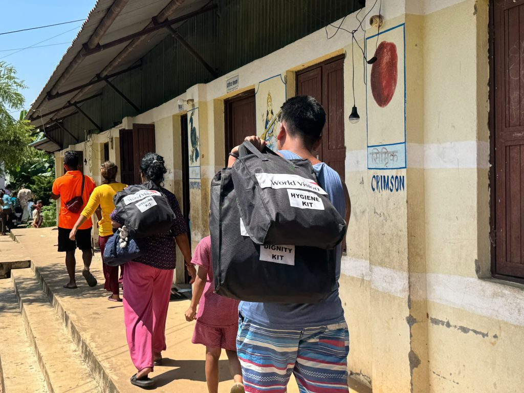 Group of people in line with World Vision hygiene kit backpacks walking beside a muraled building on a sunny day.