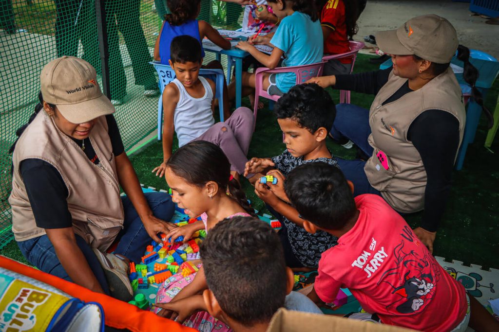 Volunteer women and kids sit on a colorful mat, building with bright plastic blocks outdoors under a green fence.