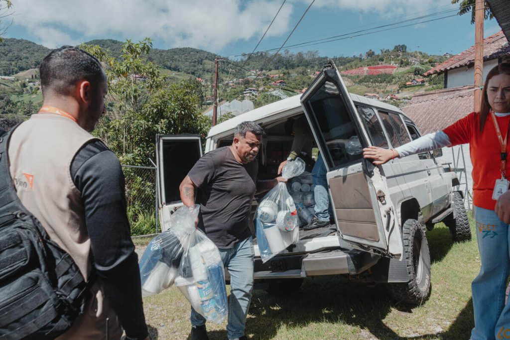 Man unloading bags of water and supplies from an open SUV in a rural hillside area. A woman in red stands nearby and a man in a vest watches over the scene.