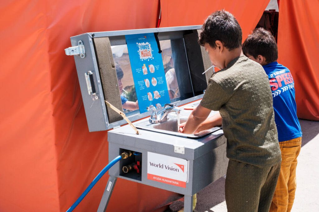 Two children at a portable handwashing station under an orange tent, washing their hands with soap and water.