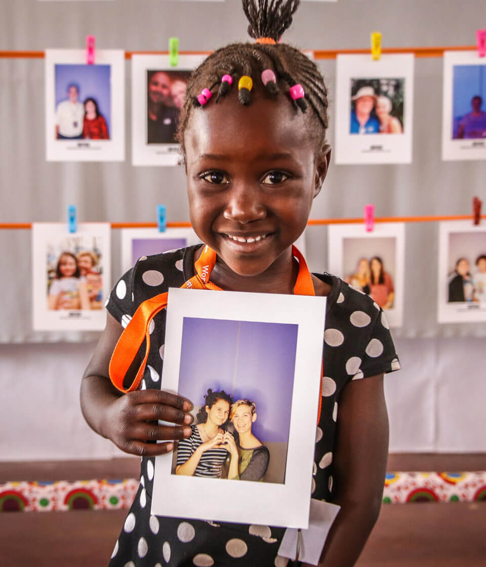 Smiling young girl with braided hair holding a framed photo of two people; colorful photo wall display behind her.