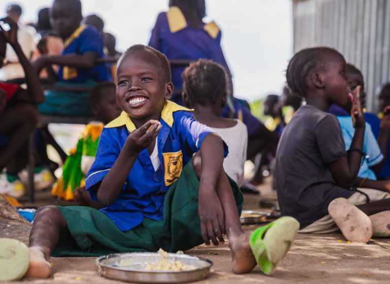 A smiling boy in a blue and yellow school uniform sits on the ground eating from a metal plate, surrounded by other children during a meal.