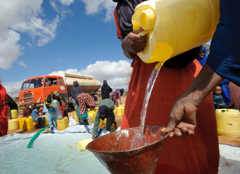 People gather at a water distribution site; a man pours water into a yellow container using a funnel, with many jerry cans nearby and a red truck in the background.