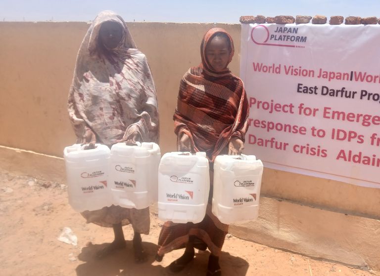 Two women in dusty robes carry white water jerrycans during aid distribution, with a relief banner in the background.