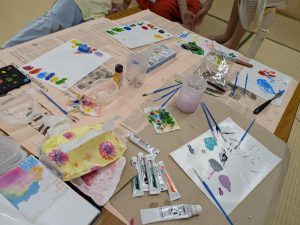 Art table covered with newspaper, showing paint tubes, brushes, water cups, and colorful painted papers in progress.